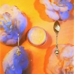 orange background with blue and white flowers surrounding a pot of skincare with a gold fork and spoon on either side
